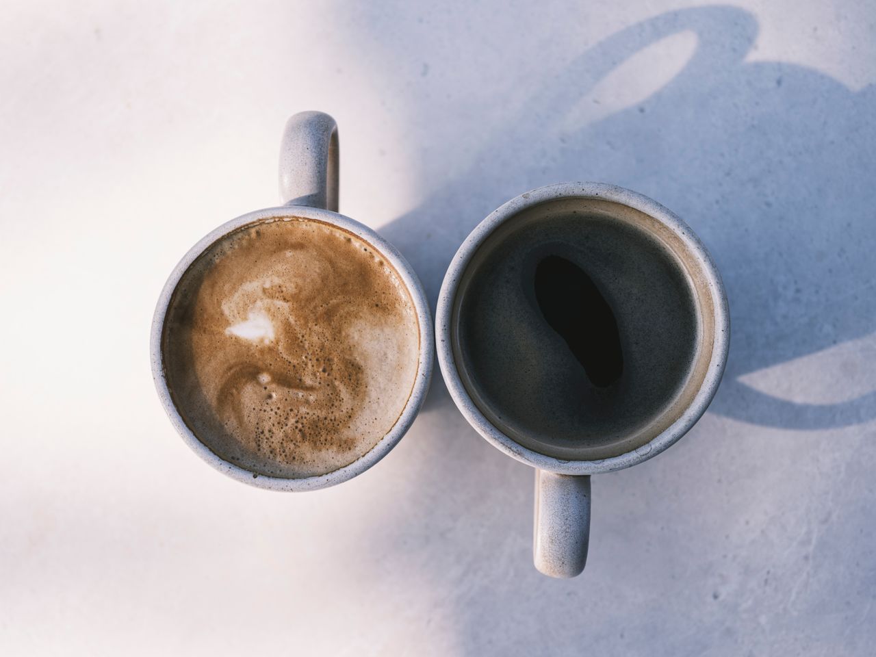 Aerial view of two coffee cups casting shadows on a sunny table, showcasing latte and black coffee.